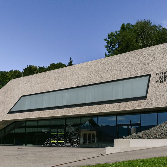 Die Vorderseite eines modernen Gebäudes mit einer großen Glaswand, auf der die Worte 'MAKE ME ARE' stehen. Rechts neben dem Gebäude befindet sich ein Haufen Steine.