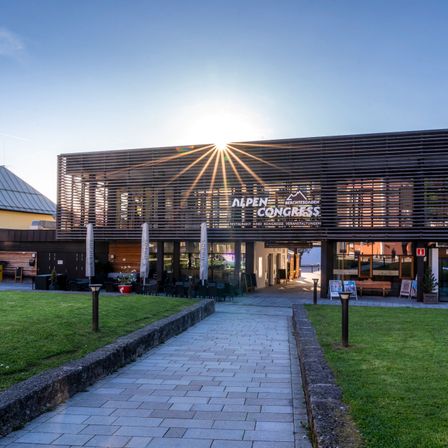 Das Alpen Congress Hotel ist modern mit Holzwänden. Eine Sonne scheint hell hinter dem Gebäude. Tische und Stühle sind draußen, mit Schirmen und einem Blumentopf.