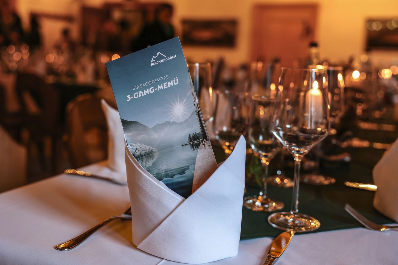 Ein Tisch mit Besteck, einer gefalteten Serviette und einem Menü für ein 3-Gänge-Menü. Gläser, eine Kerze und ein unscharfer Hintergrund.