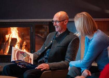 Ein älterer Mann mit Brille sitzt in einem Sessel und liest ein Buch für eine Frau neben ihm vor. Sie befinden sich in einem Raum mit einem Kamin mit Feuer dahinter.