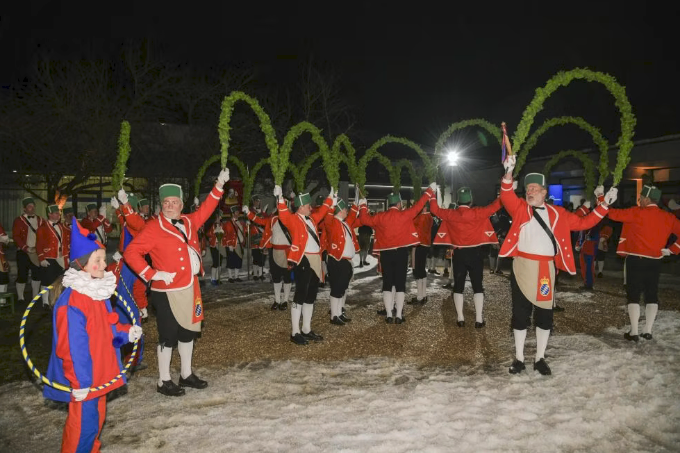 A group of people dressed in red and black costumes with green hats are standing in a snowy area at night. They are raising their hands while holding a stick. The area is surrounded by green arch-shaped structures. In the distance, there is a building with a light on.