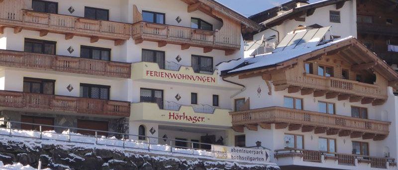 Ein mehrstöckiges Gebäude mit hölzernen Balkonen und einem Schild, das 'Ferienwohnungen' anzeigt. Schnee bedeckt den Boden und das Dach. Ein weiteres Gebäude ist rechts sichtbar, teilweise mit Schnee bedeckt.