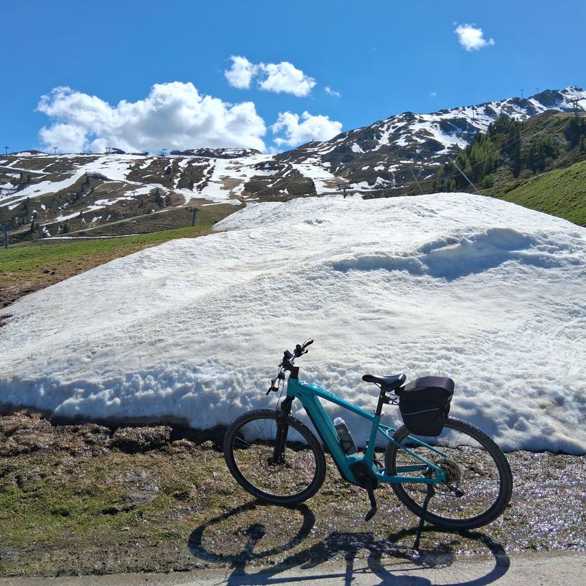 Ein blaues Fahrrad lehnt sich gegen einen schneebedeckten Hang mit Bergen im Hintergrund an einem sonnigen Tag.