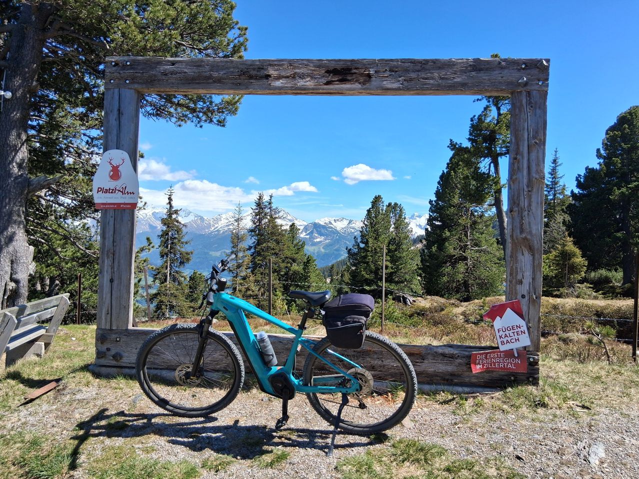 Ein blaues Elektrofahrrad steht auf einem Kiesweg neben einem Holzrahmen, mit Bergen im Hintergrund.