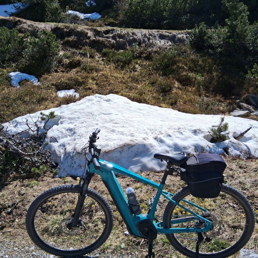 Ein türkisfarbenes Fahrrad steht an einem Hang mit schneebedecktem Bereich und Pflanzen. Eine Flasche ist am Fahrrad befestigt, und eine Tasche befindet sich auf dem hinteren Sitz.
