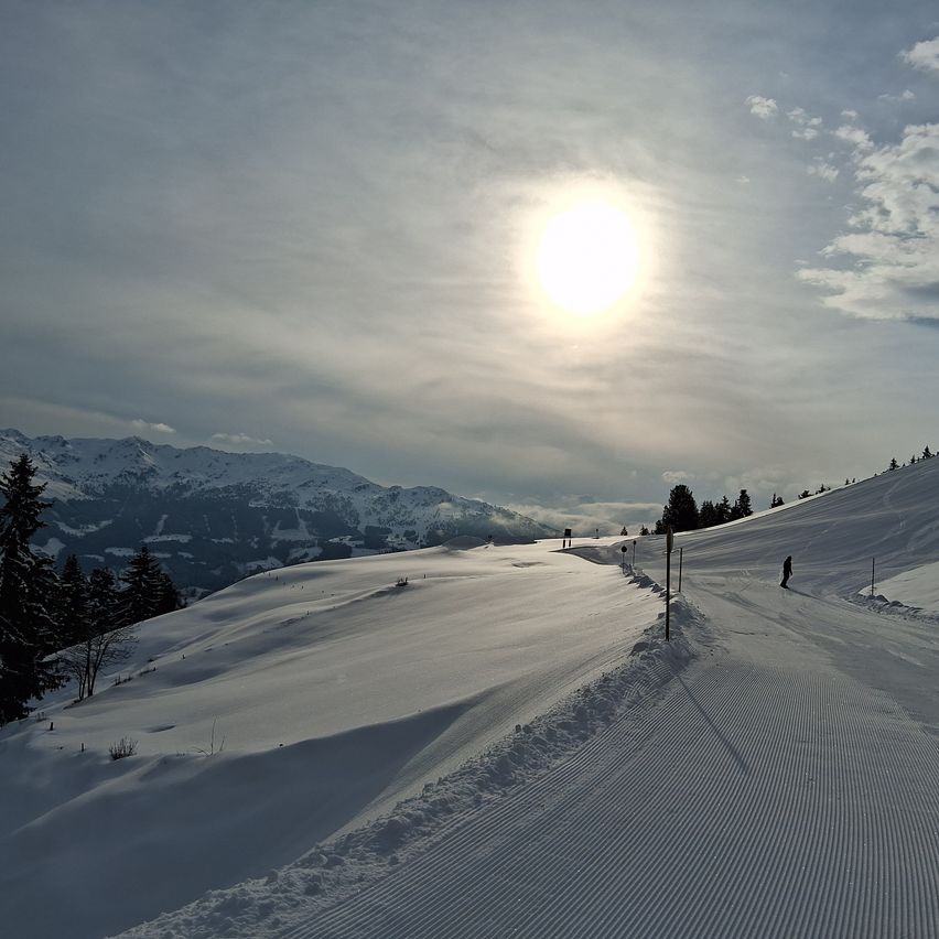 Ein sonniger Tag an einem verschneiten Berg, mit einem Skifahrer auf der Loipe und Bergen in der Ferne.