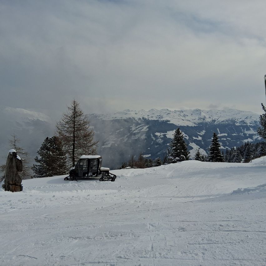 Eine schneebedeckte Berglandschaft mit einem Schneemobil und einer Holzstatue im Vordergrund. Bäume säumen die Hänge, und schneebedeckte Berge erheben sich im Hintergrund.