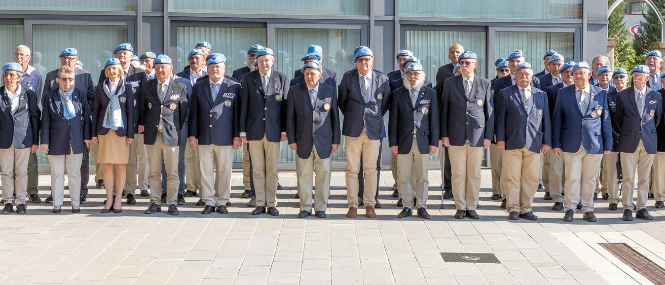 Gruppe von Veteranen in dunkelblauen Blazern mit Medaillen auf der Brust und hellblauen Mützen, die vor einem Gebäude stehen.