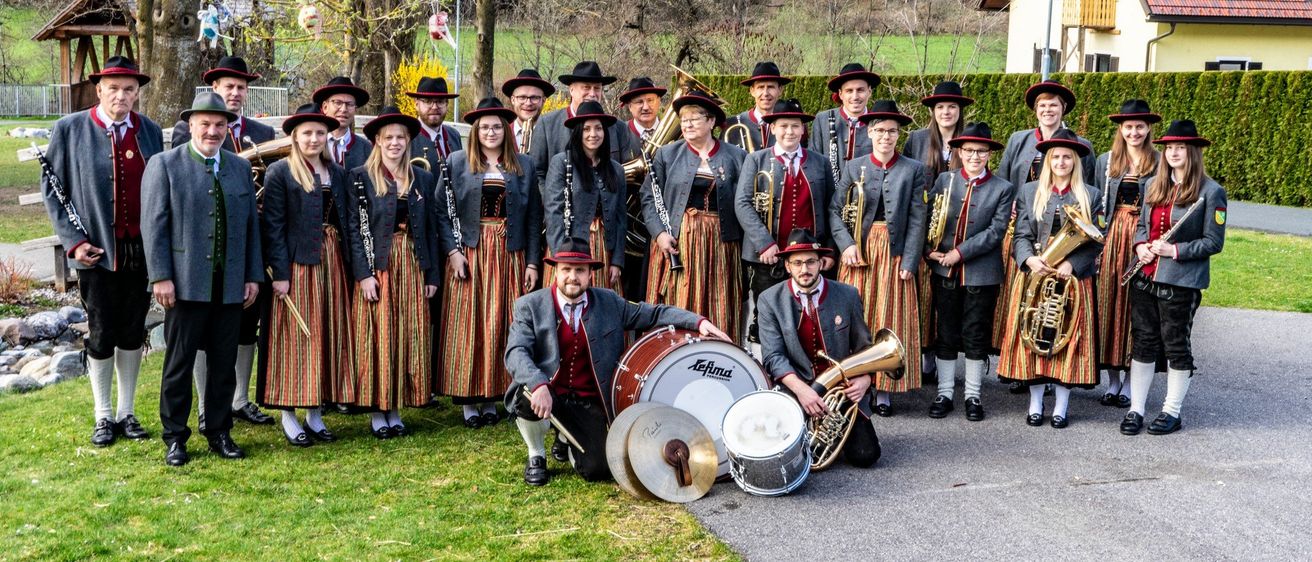 Eine Gruppe von Musikern in traditioneller Kleidung, einschließlich Hüten und Stiefeln, posiert für ein Foto. Einige halten Instrumente wie Trompeten, Tubas und Trommeln. Sie stehen vor einem Haus mit einem Zaun.