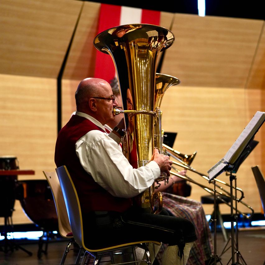 Ein Mann spielt eine Blechposaune bei einem Konzert, sitzt auf einem Stuhl auf der Bühne. Hinter ihm spielt ein weiterer Musiker eine Posaune. Im Hintergrund ist eine Holzwand mit einer Flagge und Instrumenten zu sehen.