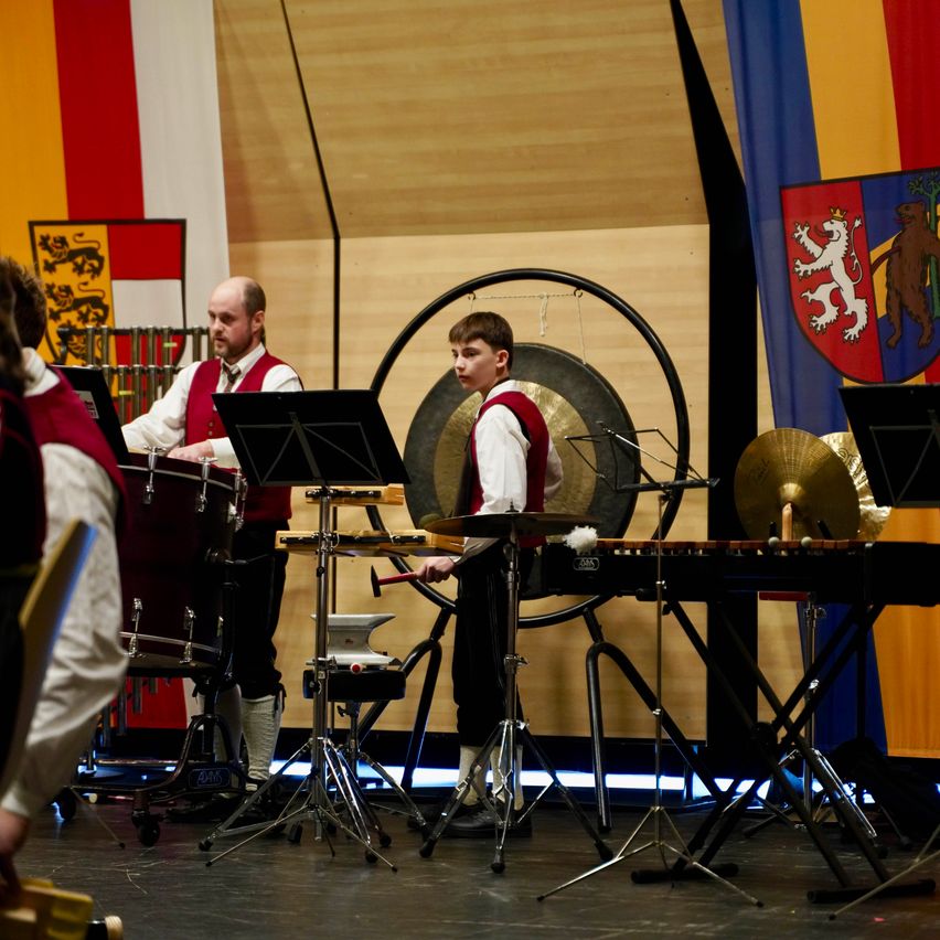 Zwei Musiker spielen Trommeln und eine Gong auf der Bühne, mit einer großen Flagge im Hintergrund.