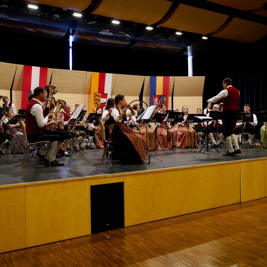 Eine Band auf der Bühne spielt mit verschiedenen Instrumenten, einschließlich Tubas und Flöten, unter Bühnenlicht. Der Dirigent steht vorne.