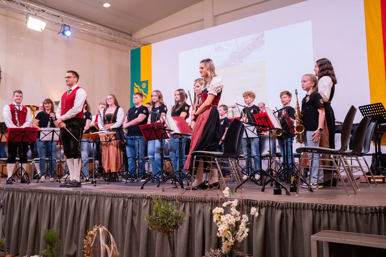 Eine Musikband tritt auf einer Bühne auf, mit einer Flagge im Hintergrund. Sie stehen vor Musikinstrumenten und einige sitzen auf Stühlen.