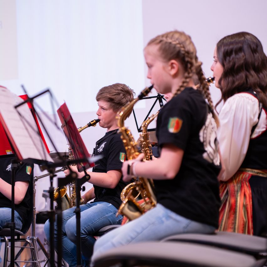 Ein Saxophon-Ensemble übt in einem Raum mit einer Projektionsleinwand. Die Gruppe besteht aus einem Jungen und drei Mädchen, die alle schwarze T-Shirts mit einem Logo tragen.
