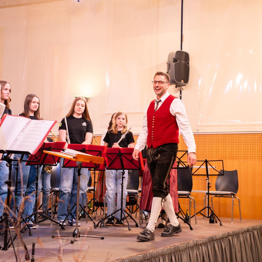 Ein Mann in einer roten Weste und schwarzen Hosen steht auf der Bühne und lächelt. Hinter ihm steht eine Gruppe von Frauen mit Musikinstrumenten.