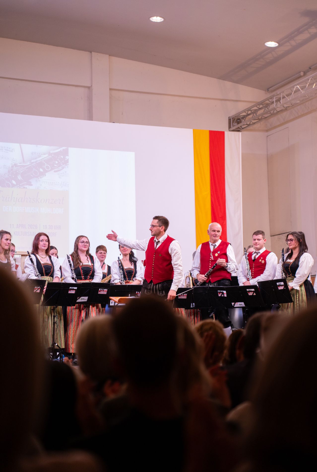 Ein Blasorchester spielt auf der Bühne, während eine Leinwand mit Noten zu sehen ist. Die Musiker sind in traditioneller Kleidung gekleidet, und ein Mann in einem roten Westchen leitet die Aufführung.