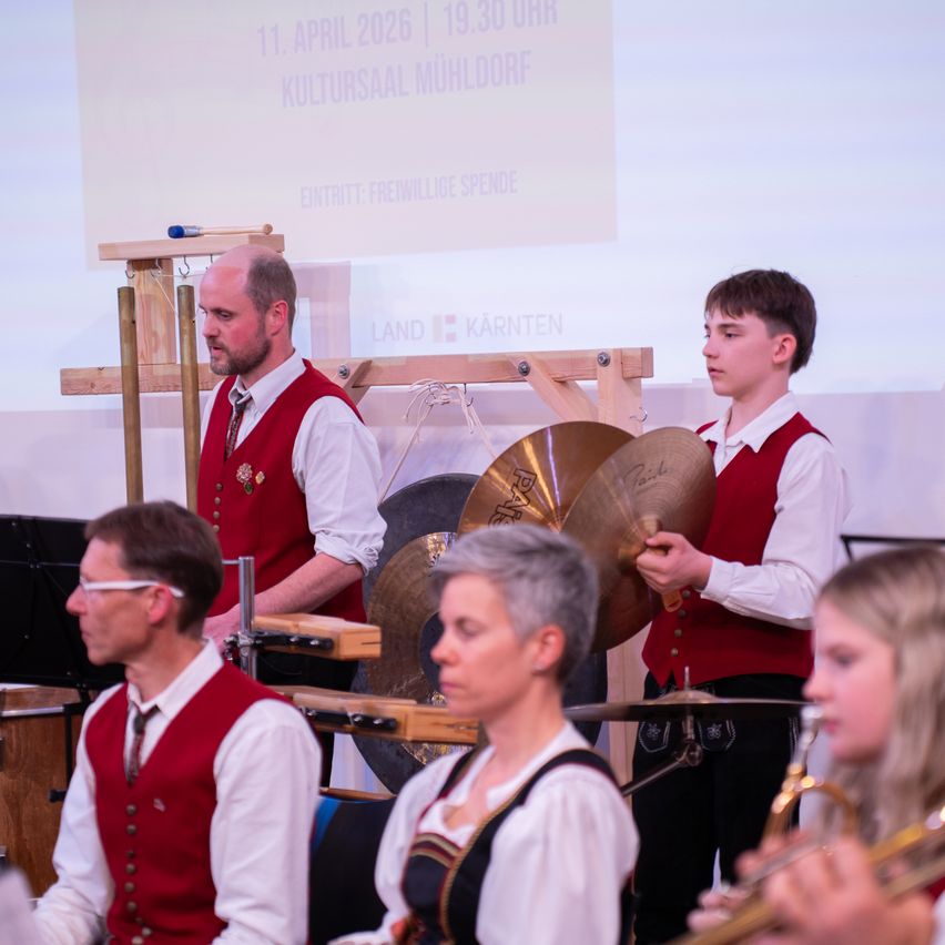 Ein musikalischer Auftritt mit verschiedenen Instrumenten, einschließlich einer Gong und einer Trompete, findet in einem Kultursaal statt. Die Veranstaltung ist für den 11. April 2026 geplant.