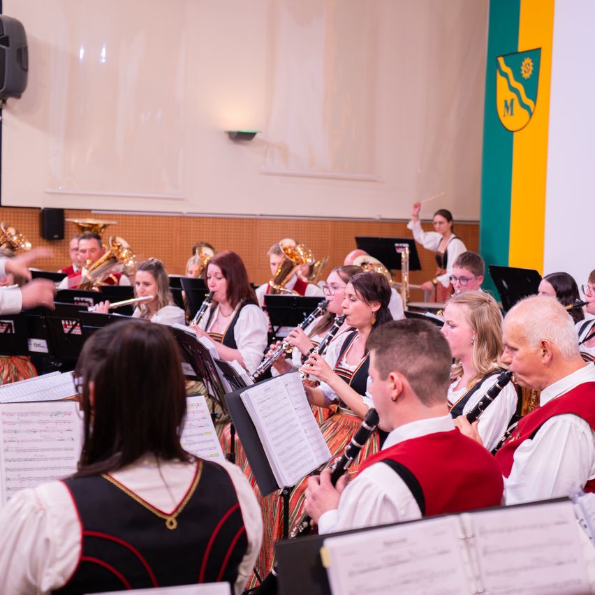 Ein Orchester spielt drinnen, mit Musikern, die verschiedene Instrumente wie Flöten, Klarinetten und Trompeten spielen. Sie sind alle in traditioneller Kleidung gekleidet. Ein Dirigent leitet die Aufführung.