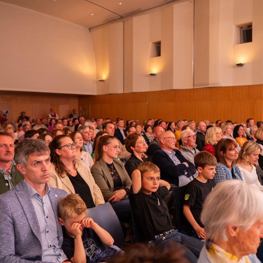 Eine große Menschenmenge, hauptsächlich Erwachsene, sitzt auf Stühlen und schaut etwas. Einige tragen Brillen, und einige haben die Arme verschränkt.