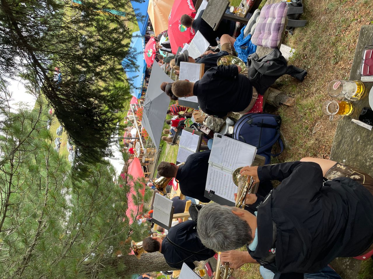 A group of musicians are practicing outdoors, some seated on the ground with music stands, while others are standing and holding brass instruments. There are tents and umbrellas in the background.