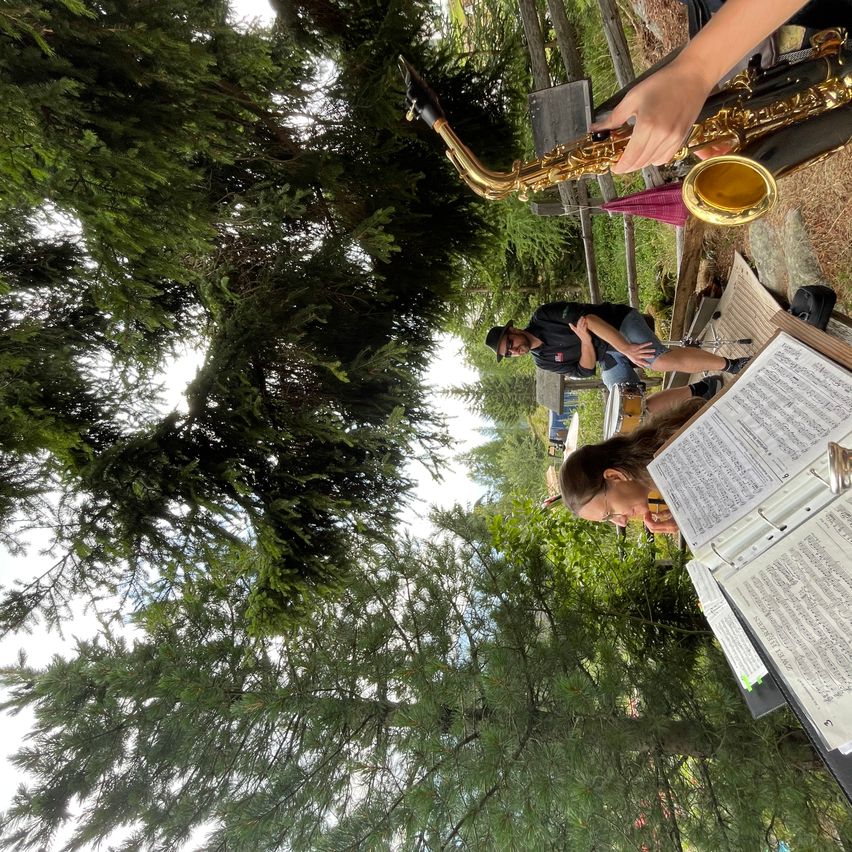 A musician plays a saxophone while another person looks at sheet music on a stand in a park.