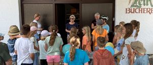 Eine Gruppe von Kindern und Erwachsenen versammelt sich vor einem Gebäude, möglicherweise einer Bibliothek, mit zwei Frauen, die vor ihnen stehen. Die Kinder halten Luftballons, und eine der Frauen trägt eine Sonnenbrille.