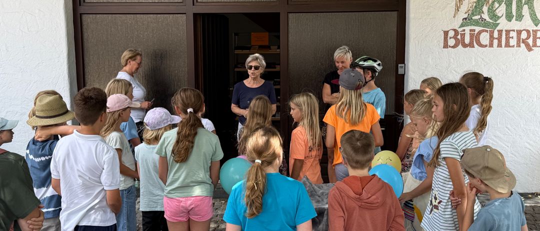 Eine Gruppe von Kindern und Erwachsenen versammelt sich vor einem Gebäude, möglicherweise einer Bibliothek, mit zwei Frauen, die vor ihnen stehen. Die Kinder halten Luftballons, und eine der Frauen trägt eine Sonnenbrille.