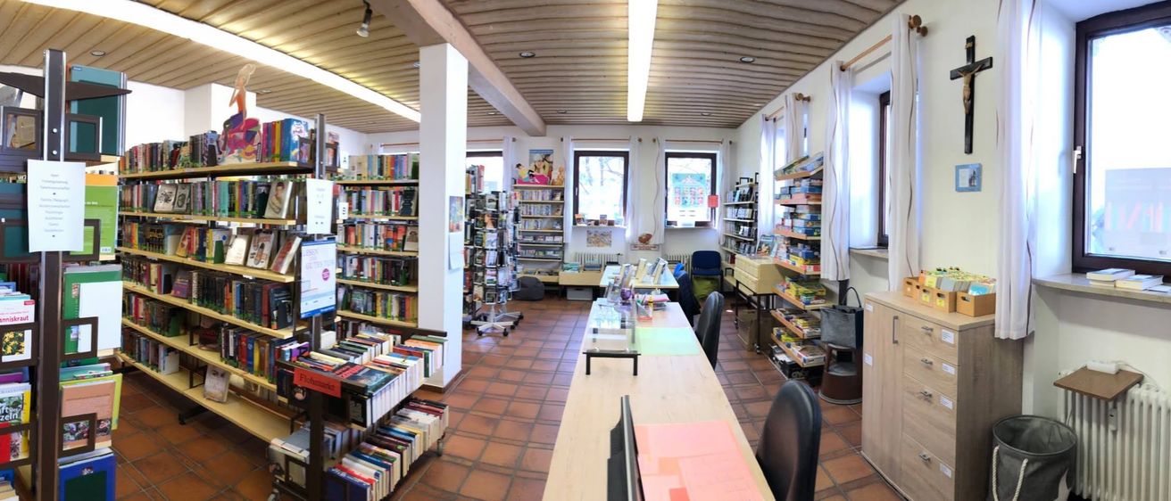 Innenansicht einer Bibliothek mit hölzernen Bücherregalen voller Bücher. Ein hölzerner Tisch mit Gegenständen darauf steht in der Mitte, umgeben von Stühlen. Ein weißer Pfeiler steht auf der linken Seite, und zwei Fenster mit weißen Vorhängen befinden sich an der Wand.
