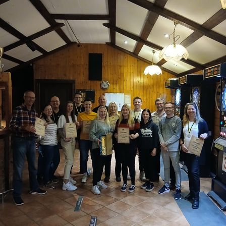 Eine Gruppe von Menschen steht zusammen in einem Raum mit Holzwänden und gefliesten Böden, hält Urkunden und lächelt für ein Foto.