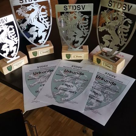 Ein Holztisch zeigt Trophäen und Urkunden. Die Trophäen haben ein Schilddesign und einen Glasbecher. Die Urkunden liegen auf dem Tisch, und der Text ist nicht klar.