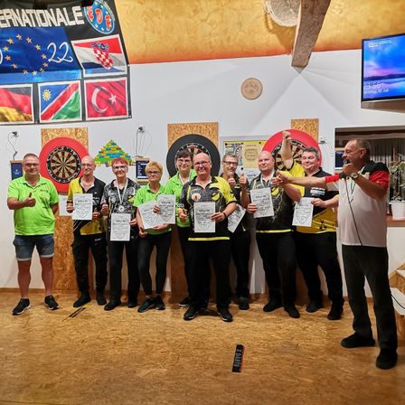 Eine Gruppe von Menschen in Grün und Gelb hält Urkunden in einem Raum mit Dartscheiben und Fahnen an der Wand. Ein Mann in Weiß hält eine Rede.