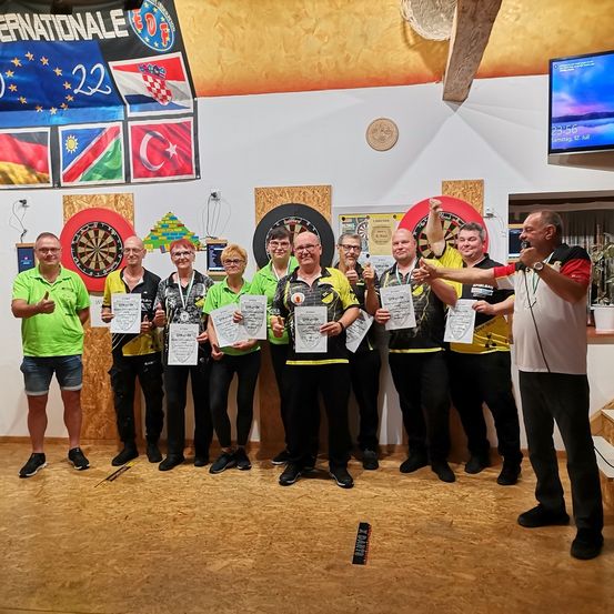 Eine Gruppe von Menschen in Grün und Gelb hält Urkunden in einem Raum mit Dartscheiben und Fahnen an der Wand. Ein Mann in Weiß hält eine Rede.