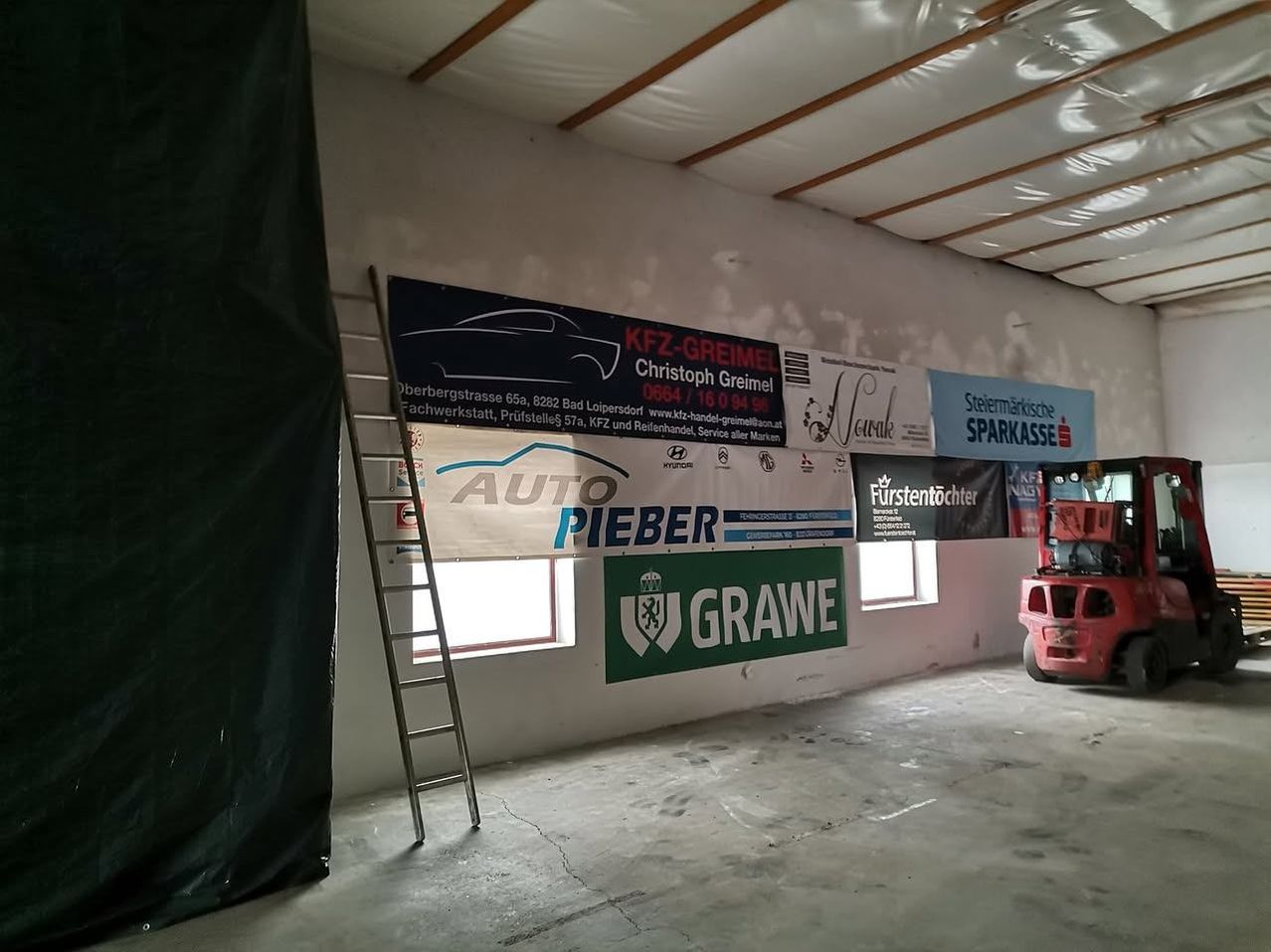 Eine leere Garage mit Bannern an der Wand und einer Leiter dagegen. Die Banner enthalten Namen und Logos. Ein roter Gabelstapler ist im Hintergrund geparkt.