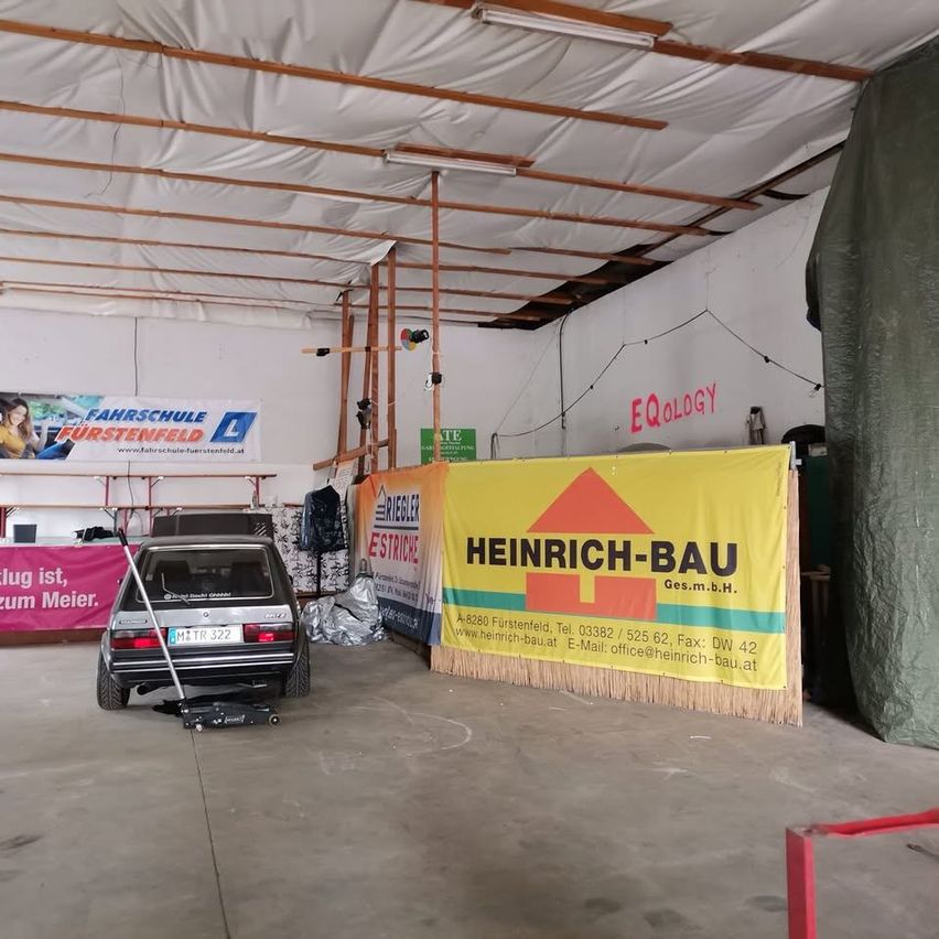 Eine Garage mit einem geparkten Auto. Ein gelbes Banner mit der Werbung Heinrich-Bau ist sichtbar. Ein weißes Banner mit dem Text Farschule Furstenfeld ist an der Wand befestigt.