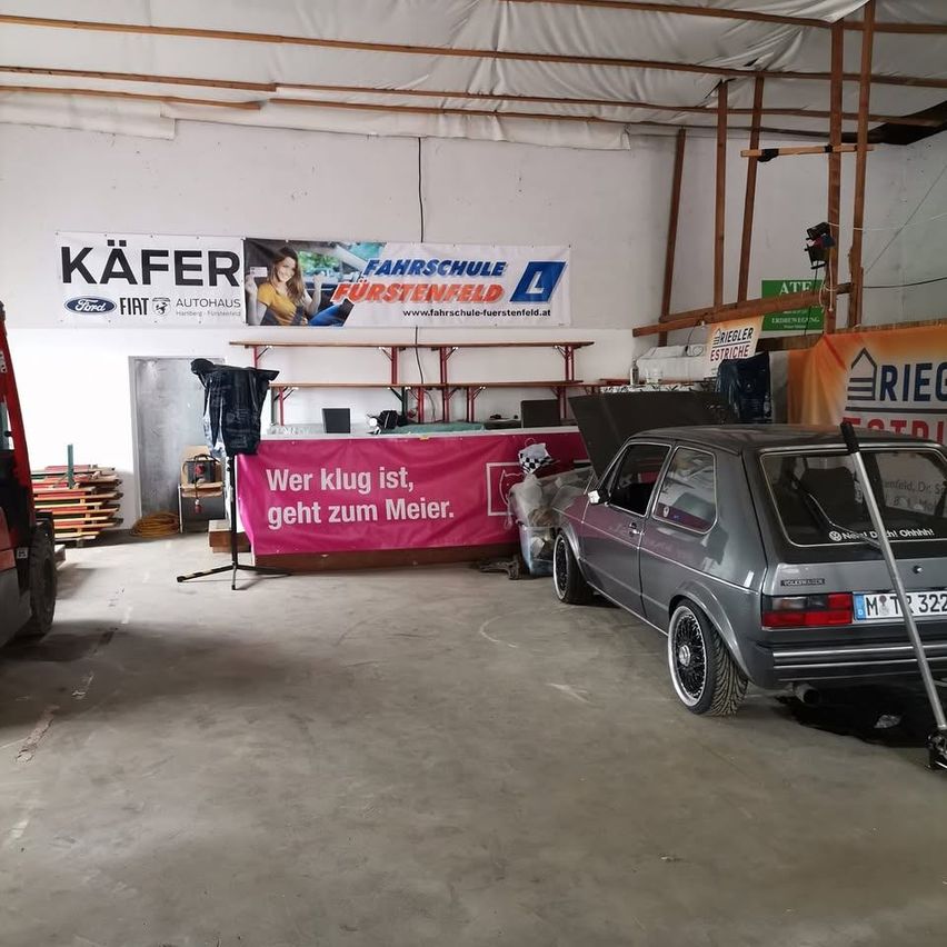 Eine Autowerkstatt mit einem Banner, das 'Wer klug ist, geht zum Meier' besagt. Ein Ford und Fiat-Schild befindet sich über dem Banner. Ein Auto ist geparkt mit offenem Motorhaube.