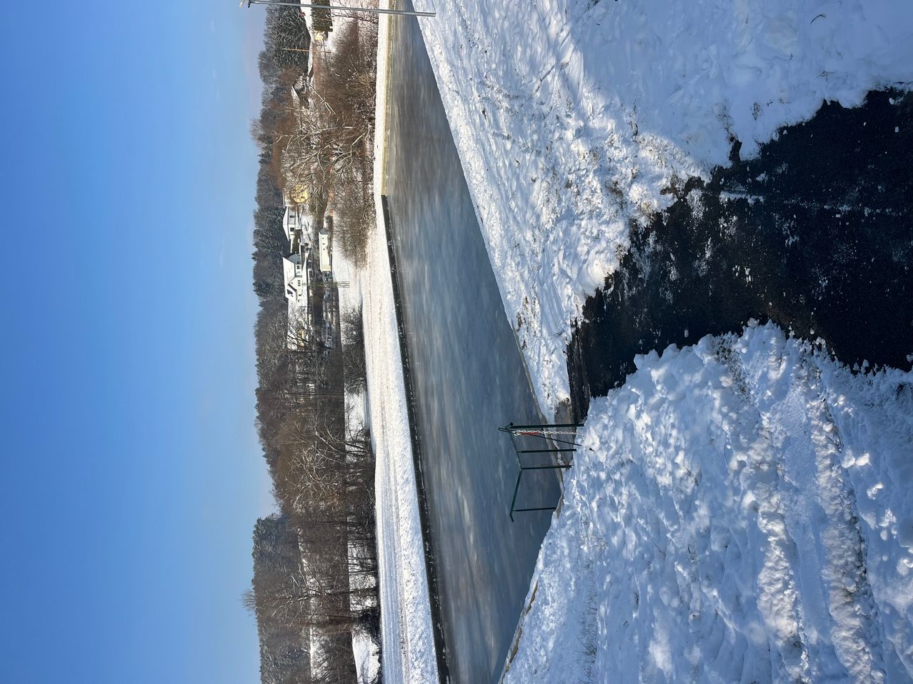 Ein gefrorener See umgeben von Bäumen und Häusern mit einem klaren blauen Himmel darüber. Schnee bedeckt den Boden und die Straße rechts.