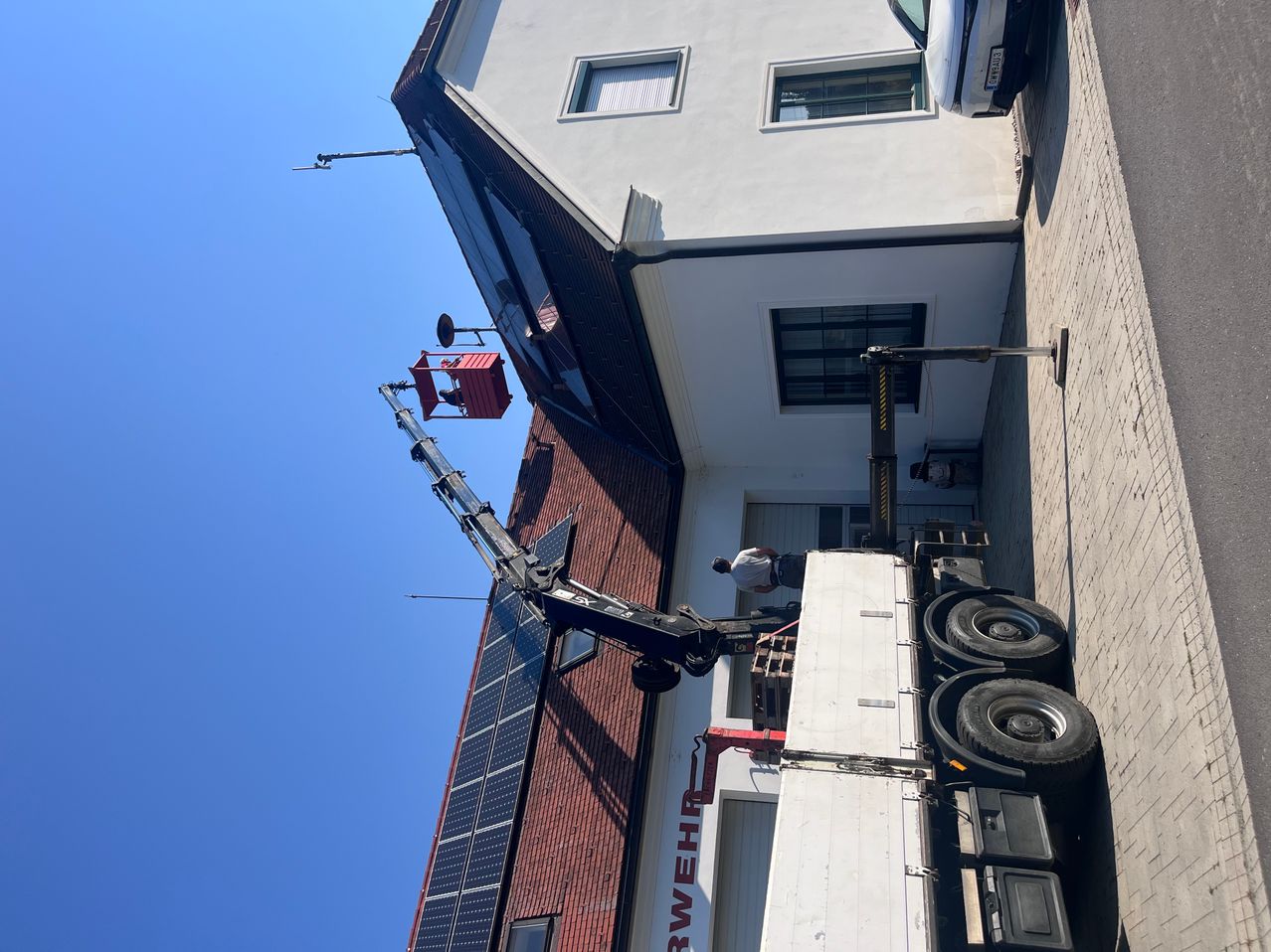 Ein LKW mit einem Kran erstreckt sich zu einem Gebäude, wo eine Person in der Nähe des Fensters arbeitet. Das Gebäude hat eine rote Ziegelwand, weiße Wände und Solarpanele an der Seite.