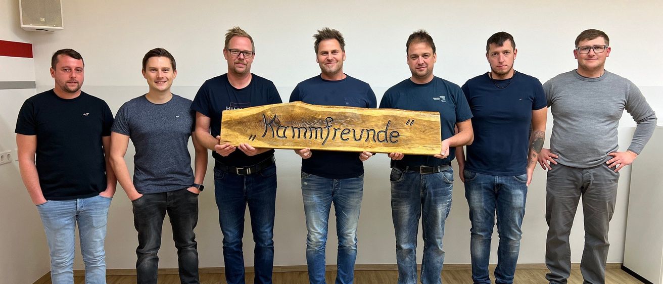 Sieben Männer stehen in einer Reihe, lächeln und halten ein Holzschild mit der Aufschrift 'Kammfreunde'. Sie sind lässig gekleidet und posieren für ein Foto. Der Ort ist ein Raum mit Holzboden und weißen Wänden.
