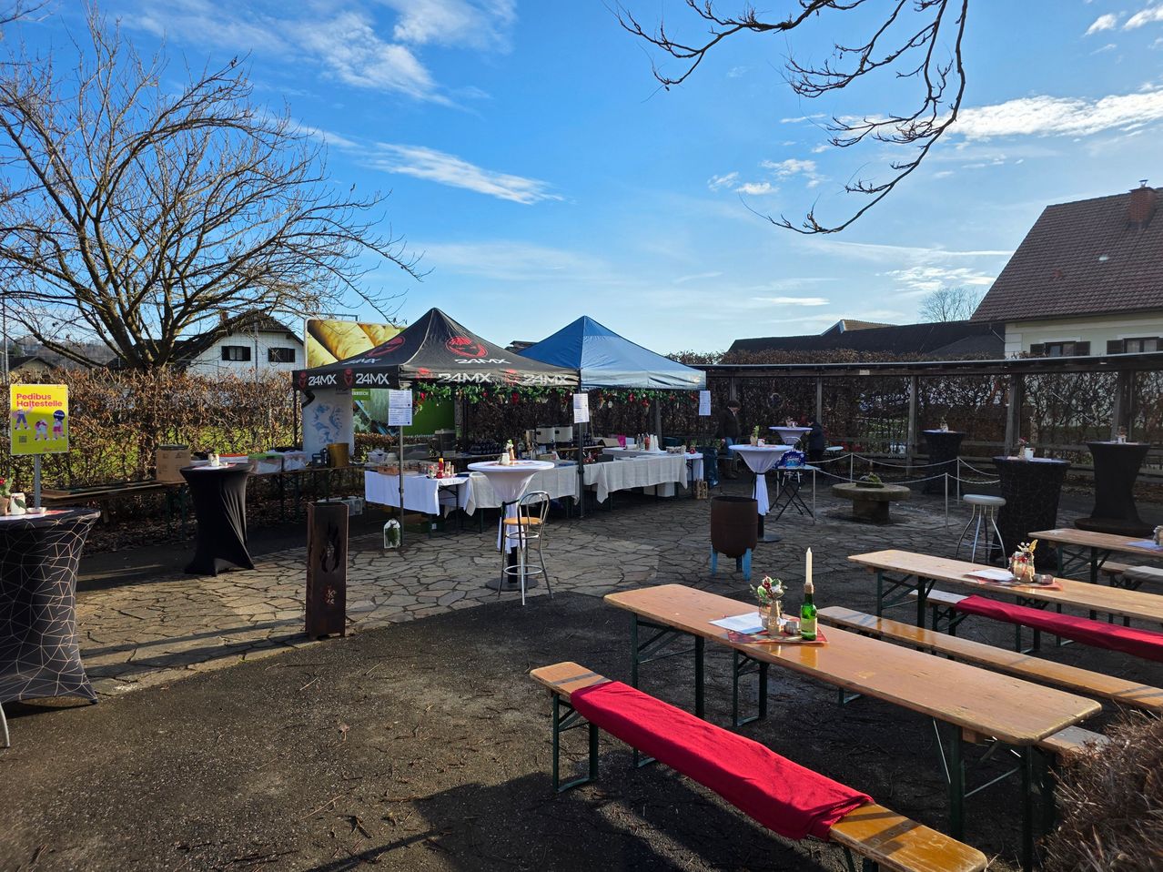 An outdoor area with multiple tables and chairs under tents. A table with a candle and a bottle is placed on a bench. Trees and houses are in the background under a clear sky.