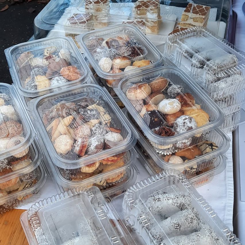 Multiple clear plastic containers filled with various pastries are arranged on a white surface. The pastries include doughnuts, cookies, and other baked goods, some dusted with powdered sugar.