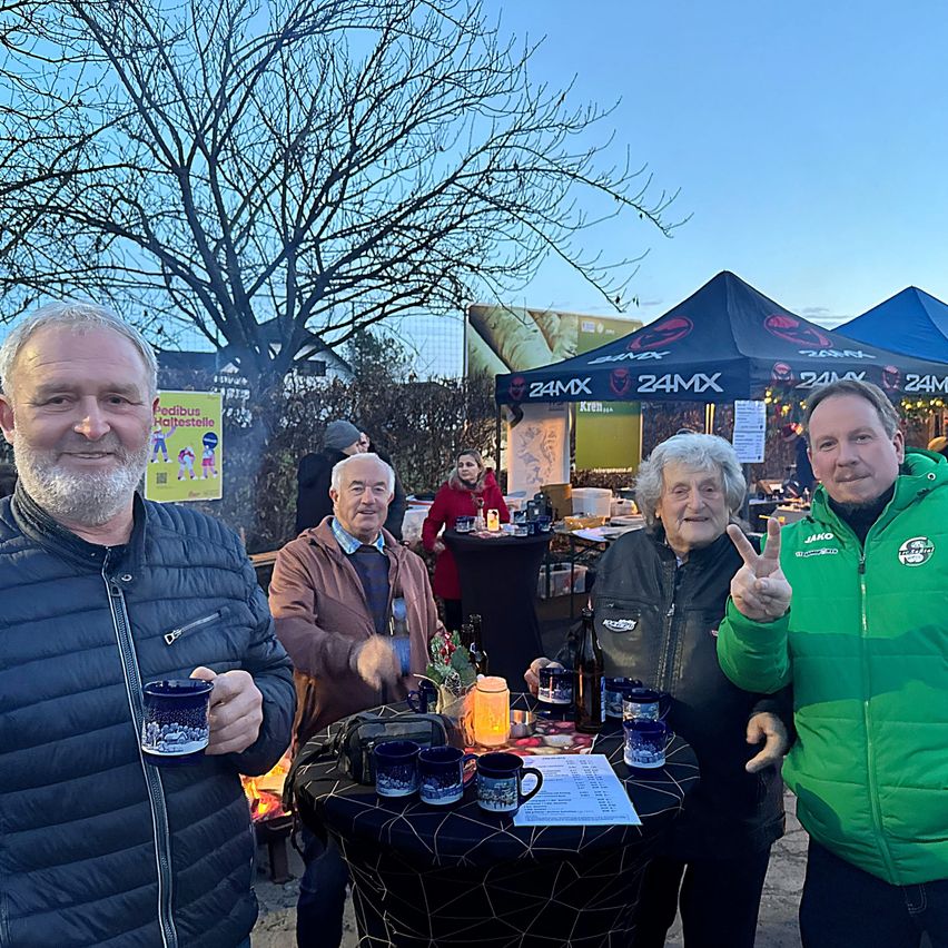 Four people stand outside near a table with mugs and a sign, one man makes a peace sign. In the background, tents are set up with signs and people.