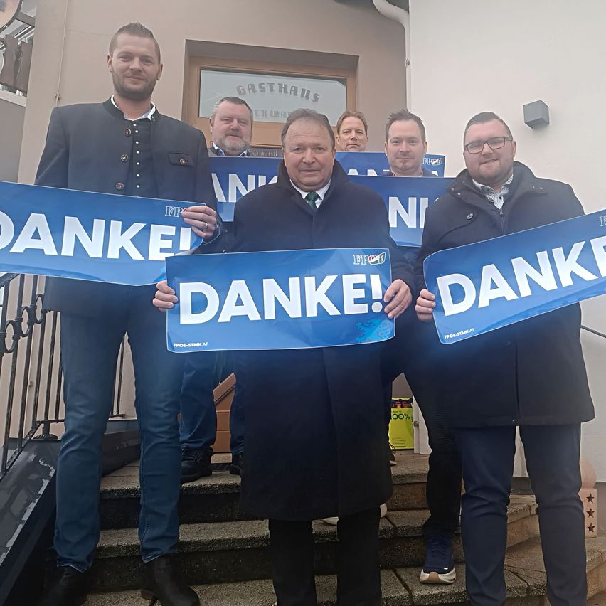 Fünf Männer in Anzughose stehen auf einer Treppe und halten blaue Banner mit dem Wort 'DANKE!'. Der Mann in der Mitte trägt einen Krawatte und hält ein Banner in beiden Händen. Das Gebäude hat ein Schild mit dem Text 'Gasthaus'.