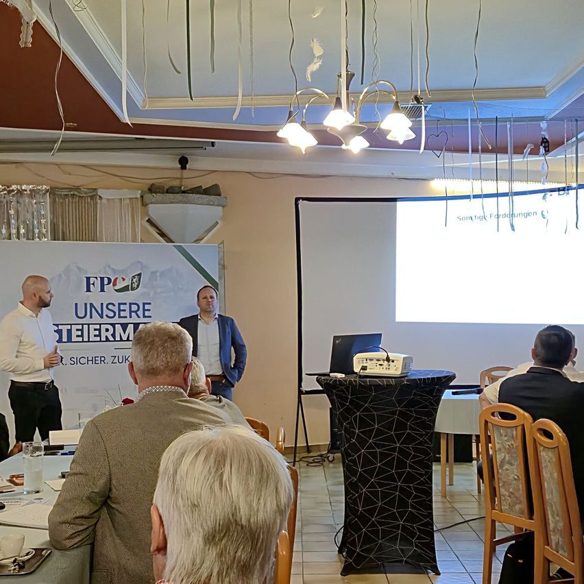 A man is giving a presentation with a projector on a podium in a conference room. A large banner with a logo and text is behind him.