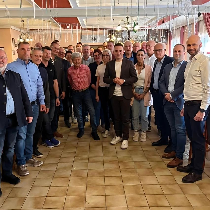 A group of men and women in formal attire stand together in a room, posing for a photo. The room has a tiled floor and a ceiling with hanging decorations.