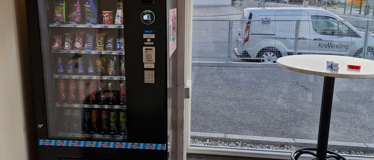 Ein schwarzer Verkaufsautomat mit Snacks und Getränken steht in einem Raum mit offener Tür. Draußen ist ein weißer Lieferwagen am Straßenrand geparkt, mit Häusern in der Ferne.