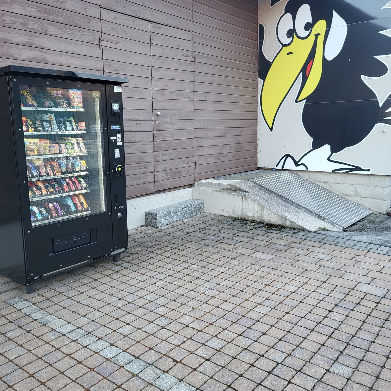 Ein Verkaufsautomat voller Snacks steht neben einem Gebäude mit einer Vogelmalerei. In der Nähe führt eine Rampe mit einem Metallgitter zum Eingang.