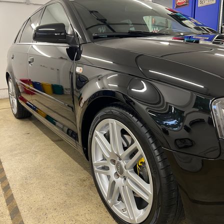 Ein schwarzer Wagen mit poliertem Äußeren steht in einer Garage. Der Wagen hat ein schlankes Design mit glänzendem Finish. Das Vorderrad hat silberne Leichtmetallfelgen.