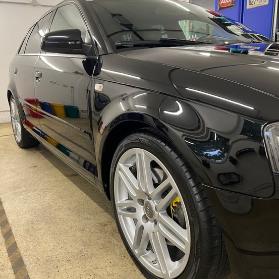 Ein schwarzer Wagen mit poliertem Äußeren steht in einer Garage. Der Wagen hat ein schlankes Design mit glänzendem Finish. Das Vorderrad hat silberne Leichtmetallfelgen.
