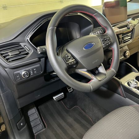 Innenraum eines Ford-Autos mit Lenkrad und Bedienfeld. Das Lenkrad hat ein Ford-Logo und Tasten. Das Bedienfeld hat einen Gangschalter und Klimareglertasten. Der Boden hat Teppichmatten.