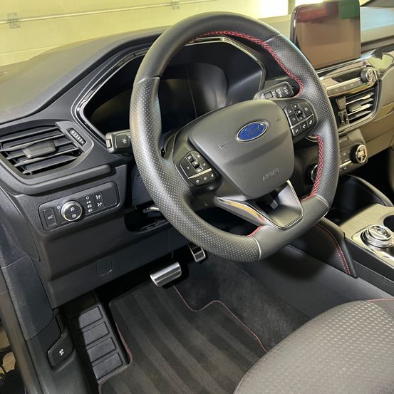 Innenraum eines Ford-Autos mit Lenkrad und Bedienfeld. Das Lenkrad hat ein Ford-Logo und Tasten. Das Bedienfeld hat einen Gangschalter und Klimareglertasten. Der Boden hat Teppichmatten.